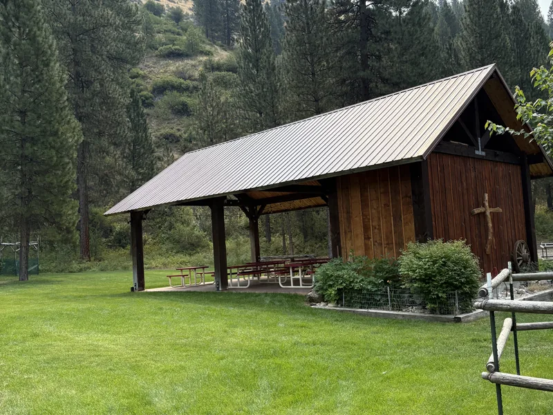 The timber chapel building beside the covered pavilion, with the mown lawn in front and a wooded ridge behind.