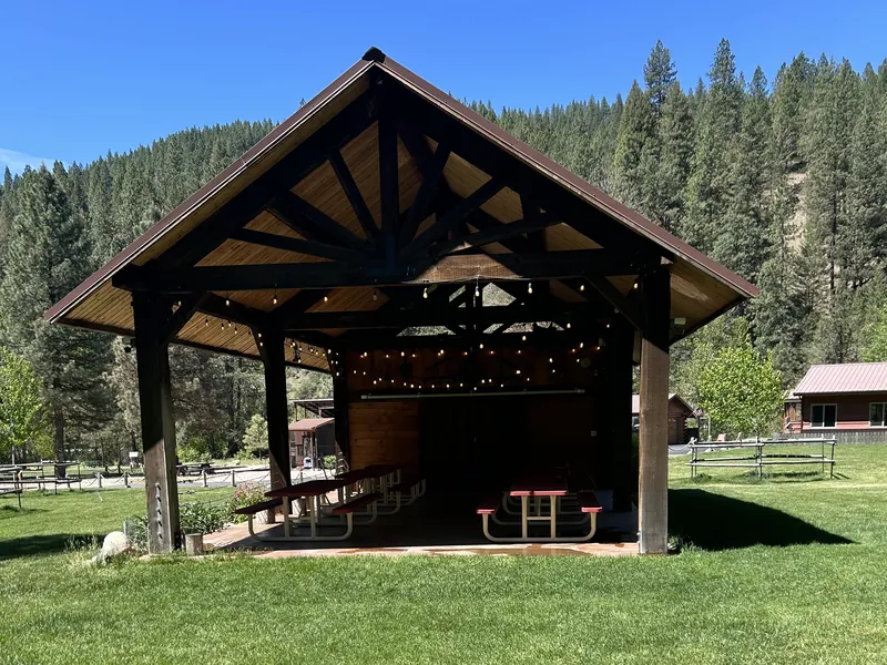 The open-air timber pavilion strung with lights, tables set inside and pines rising behind.